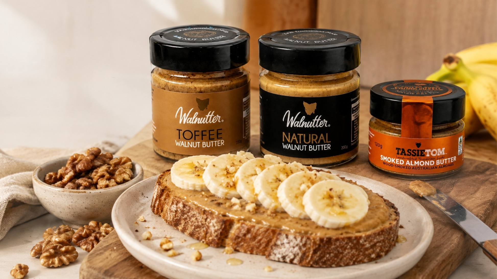 Walnututter products including jars of toffee walnut butter and natural walnut butter, with a plate of toast topped with banana slices on a wooden table.