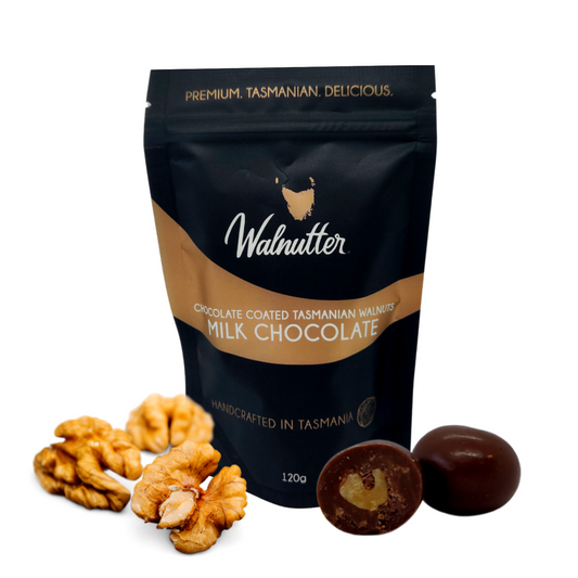 Premium. Tasmanian. Delicious. Walnutter Tasmanian premium walnuts coated in Milk Chocolate - Handcrafted in Tasmania - 120g black and gold foil pouch bag, with fresh walnuts halves and chocolates sitting next to the bag.